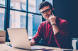 © BullRun - Young positive businessman calling to colleague via application on cellphone while sitting at desktop with laptop, portrait of cheerful hipster guy in eyewear searching information for startup project