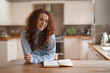 © Iryna - Young woman with curly hair writes in a notebook in a cozy kitchen during a sunny afternoon