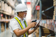 © KANGWANS - A man in a white shirt and yellow vest is using a tablet to scan a barcode. He is wearing a hard hat and safety glasses, indicating that he is working in a warehouse or industrial setting