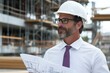 © Milos - A male engineer in formal attire and a helmet is intently reviewing construction plans on a busy site, showcasing professionalism and dedication to his work.