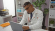 © Krakenimages.com - Hispanic male doctor working on a laptop in a clinic, managing phone call and looking focused in an indoor medical office setting.