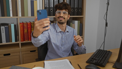 Wall Mural - Young hispanic man takes a selfie in an office while sitting at a desk with documents bookshelves and a computer in the background