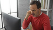 © Krakenimages.com - Hispanic man in a red sweater focused on a computer screen in an office setting, emphasizing thoughtful concentration and workplace ambiance.