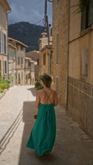 Wall Mural - Young woman in green dress exploring the charming narrow streets of valldemossa, a picturesque village in mallorca, spain, surrounded by rustic stone buildings and mountains.