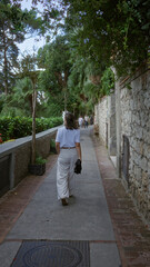 Wall Mural - Young woman walking through charming capri streets, surrounded by lush greenery and rustic stone walls under a clear sky, capturing serene italian outdoor beauty.