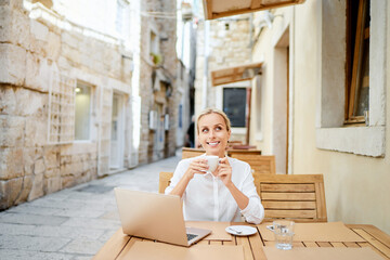 Wall Mural - Technology and travel. Working outdoors. Freelance concept. Pretty young woman using laptop and drinking cofee in sidewalk cafe on ancient europian street.