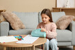 © Pixel-Shot - Cute little happy girl with pencil case, notebooks and markers sitting on chair at home