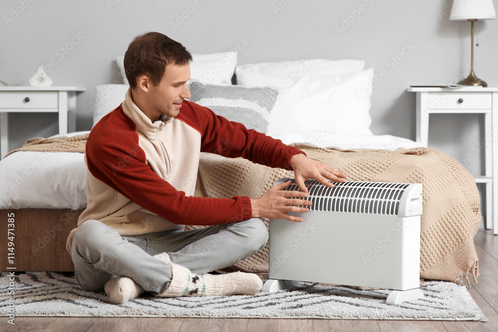 Young man warming hands near electric convector heater in bedroom