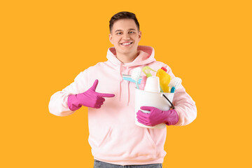 Wall Mural - Young man pointing at bucket of cleaning supplies on yellow background