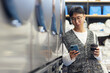 © Seventyfour - Medium shot of young Asian man with cup of coffee in hand text messaging on phone while waiting in coin laundromat standing next to line of washing machines, copy space