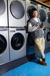 © Seventyfour - Full length shot of relaxed Asian man standing next to tumbling washing machines text messaging on phone while waiting in coin laundromat, camera flash
