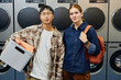 © Seventyfour - Portrait shot of young woman and Asian man with basket of clothes posing against washing machines in self service laundromat while doing household chores together
