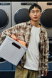 © Seventyfour - Vertical shot of young Asian man carrying basket with clothes preparing to load washing machine while confidently looking at camera in self service laundry