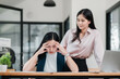 © Satori Studio - A businesswoman experiencing stress at her desk while a colleague offers support in a contemporary office setting.