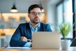 © Vilaysack - A focused man in a suit sits at a desk, gazing thoughtfully at a laptop in a modern office setting with plants and warm lighting.