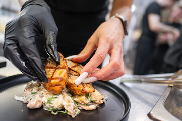 Wall Mural - A chef's hand, wearing a black glove, carefully arranges grilled chicken pieces on a plate with creamy mushroom sauce. The kitchen setting is busy and vibrant, showcasing culinary artistry.