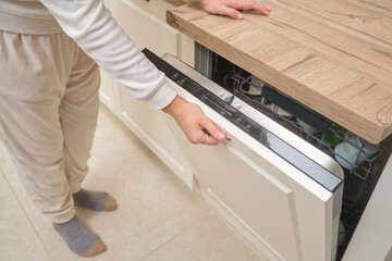 Person opening dishwasher in a modern kitchen with wooden countertop. Home appliance usage and daily kitchen routine concept.