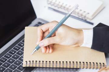 Poster - A woman's hand seen in close-up, writing in a notepad on a contemporary laptop, against a soft-focus backdrop