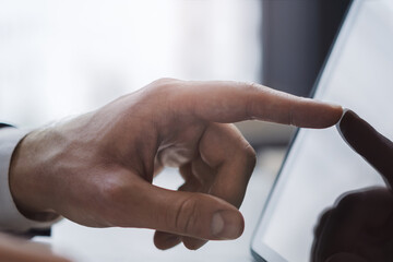 Aufkleber - Close detail of a man's finger making contact with the screen of a sleek digital tablet resting on a desk, set against the soft-focus backdrop of a light-filled office
