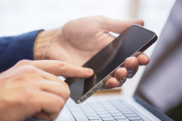 Wall Mural - Detailed view of a man utilizing his modern smartphone at his workspace, laptop in the vicinity against the fuzzy backdrop of his busy office environment