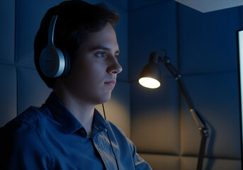 Poster - Young man concentrating on his computer screen late at night, wearing headphones and working under a desk lamp