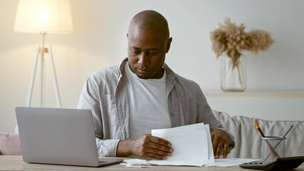 Poster - A focused African American man in his 30s reviews paperwork while sitting at a desk with a laptop in a cozy home office. Soft light adds to the peaceful atmosphere of productivity.