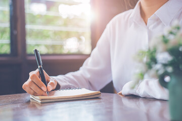 Wall Mural - Close-up a woman's hand writing on a notepad with a black pen on a wooden desk in a window-side office.