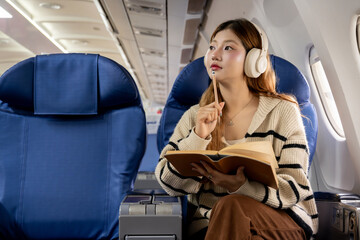 Fototapete - Asian woman sitting on airplane listening to music and writing diary while traveling somewhere during vacation.