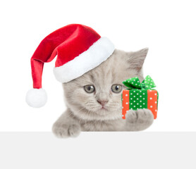 Poster - Cute tiny  cat wearing red santa hat holding gift box and looking above blank white banner. isolated on white background