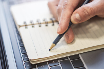 Poster - Closeup illustration of a man's hand engaged in writing in a notepad on a stylish laptop, with a soft blur in the background