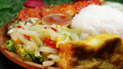 Wall Mural - Close up a plate of warm white rice served with fried fish in spicy red sambal, fried tofu, fried tempeh, fresh sautéed vegetables, shrimp paste sambal, cucumber slices, and other fresh sides.