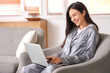 © Pixel-Shot - Young woman in grey pajamas working with laptop on armchair at home