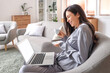 © Pixel-Shot - Young woman in grey pajamas with coffee and laptop working on armchair at home