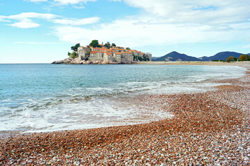 Scenic Mediterranean beaches: landscape with the Adriatic Sea, a red pebble beach and an island with old stone houses in the background. View of the resort of Sveti Stefan, Montenegro