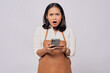 © Bangun Stock Photo - Shocked young Asian woman barista barman employee wearing brown apron working in coffee shop using mobile phone isolated on white background. Small business startup concept