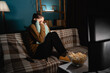 © Andrii Lysenko - Frightened 30s woman watching a horror movie on TV, holding pillow on sofa in the living room at night