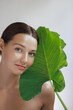 © Buyanskyy Production - A young woman with glowing, healthy skin poses beside a large green leaf, evoking a natural beauty and wellness theme. Ideal for promoting skincare, eco-friendly products, and wellness concepts that