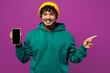 © ViDi Studio - Young Indian man in green hoody yellow hat casual clothes hold in hand use mobile cell phone with blank screen workspace area point finger aside isolated on plain purple background Lifestyle concept