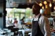 © nsit0108 - Thoughtful waiter wearing apron standing in restaurant dining room with blurred customers in background