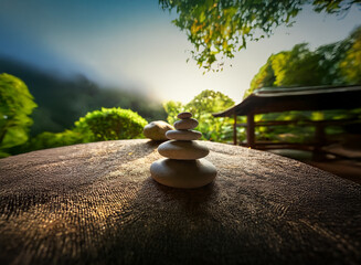 Aufkleber - Serene garden scene at sunrise.  Balanced stones on a textured surface,  peaceful ambiance with misty mountains and lush foliage in the background. A wooden gazebo is visible.