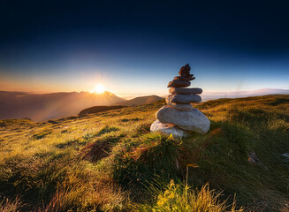Poster - Serene sunrise over majestic mountains. A carefully balanced rock cairn stands in a vibrant meadow, bathed in the golden light.  Peaceful, tranquil atmosphere.