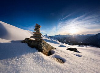 Poster - A serene winter landscape showcasing a carefully balanced rock cairn atop a snow-covered mountain.  The sun sets behind a majestic mountain range, casting a warm glow on the scene.
