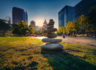 Poster - A carefully balanced rock cairn sits in a vibrant urban park at sunset, modern skyscrapers forming a backdrop.  The scene evokes serenity amidst the city's energy.