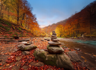 Poster - Serene autumn scene: a balanced rock cairn by a river, surrounded by vibrant fall foliage and misty mountains.  The colors are rich and warm, evoking a sense of peace and tranquility.