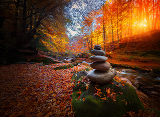 Fototapete - Serene autumn scene: a balanced stone stack beside a flowing stream, nestled amongst vibrant fall foliage.  Sunlit forest creates a magical ambiance.