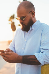 Fototapete - Handsome man in stylish outfit checking mail on smartphone app confirming booking on web page standing outdoors, bearded hipster guy blogging in social networks share files via modern mobile phone