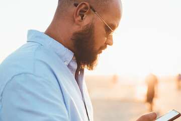 Fototapete - Young bearded male holding smartphone browsing information about cafes spending time on street, handsome hipster guy watching online video in social networks using mobile telephone during free time