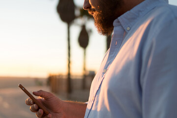 Fototapete - Cropped view of hipster guy holding modern mobile phone in hand reading news from browsed web page, young man sending text message on smartphone using 4G internet connection standing outdoors
