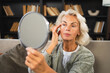© Юлия Завалишина - Love yourself. Beautiful old mature woman enjoying mirror reflection. Senior adult older lady looking at mirror confident happy on sofa at home. Woman doing daily morning beauty routine. Self love