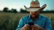 © Dulemegapixel - A man wearing a cowboy hat and clothes gazes down at his smartphone in a vast, open field, creating a blend of tradition and modernity in the serene landscape.